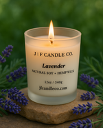 A lit “Lavender” candle by J | F Candle Co., made with natural soy and a hemp wick, is displayed outdoors on a stone surface surrounded by fresh lavender sprigs and green foliage, evoking a calm, botanical setting.