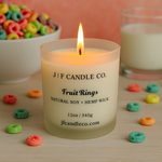 A lit “Fruit Rings” candle by J | F Candle Co., made with natural soy and a hemp wick, sits surrounded by colorful cereal loops on a light wooden surface, with a bowl of cereal and a glass of milk in the background.