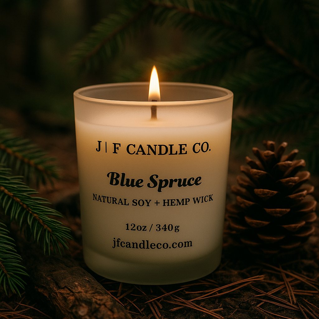 A Blue Spruce scented candle by J | F Candle Co. sits outdoors on a forest floor surrounded by pine needles, moss, and bark. The candle is lit with a small, natural flame, casting a warm glow against the wintry woodland setting.