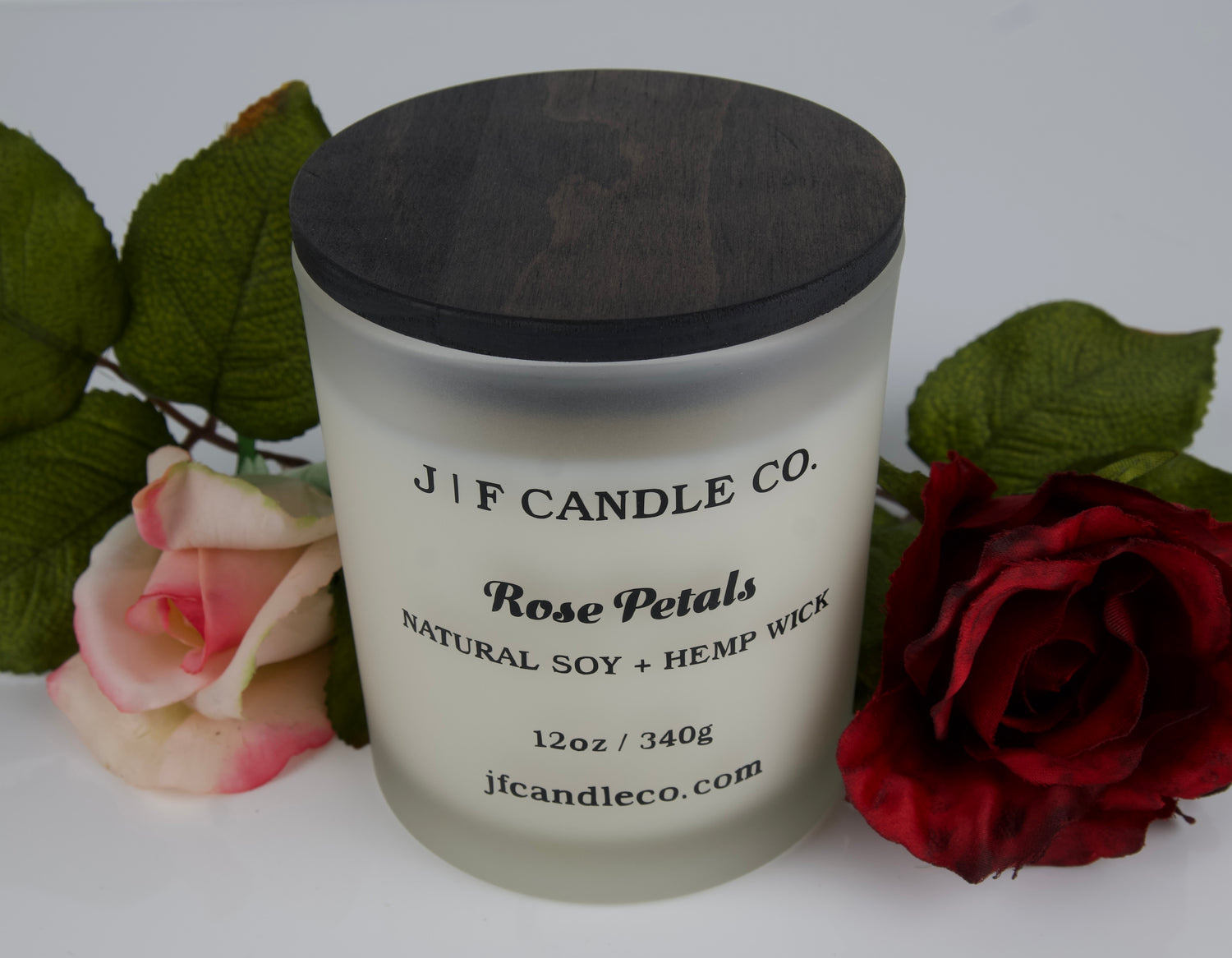 A frosted glass candle with a dark wooden lid is labeled "J | F Candle Co. Rose Petals, Natural Soy + Hemp Wick, 12oz / 340g, jfcandleco.com." The candle is displayed with red and pink roses and green leaves, creating a romantic and elegant presentation.