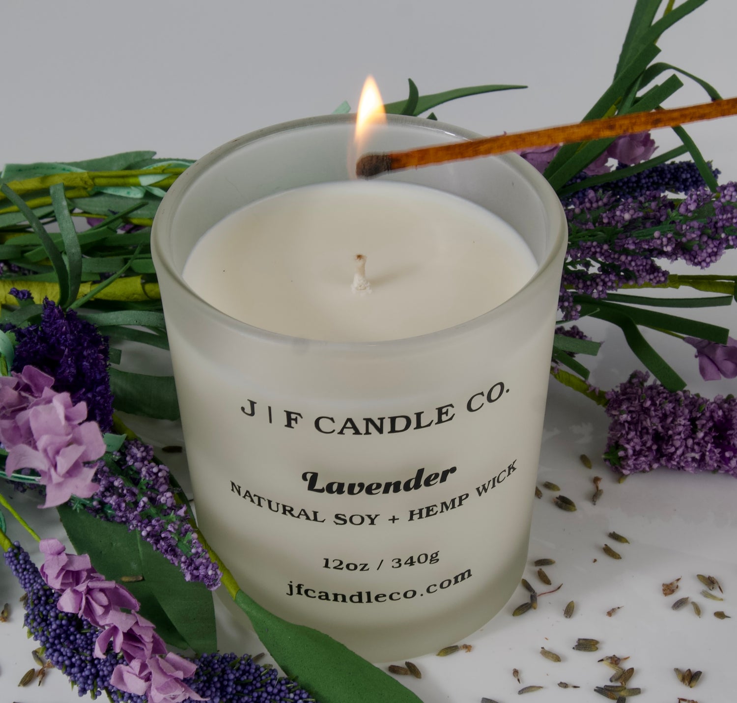 A frosted glass candle labeled "J | F Candle Co. Lavender, Natural Soy + Hemp Wick, 12oz / 340g, jfcandleco.com" is being lit with a match. The candle is surrounded by sprigs of fresh and dried lavender, adding a natural, calming aesthetic to the scene.