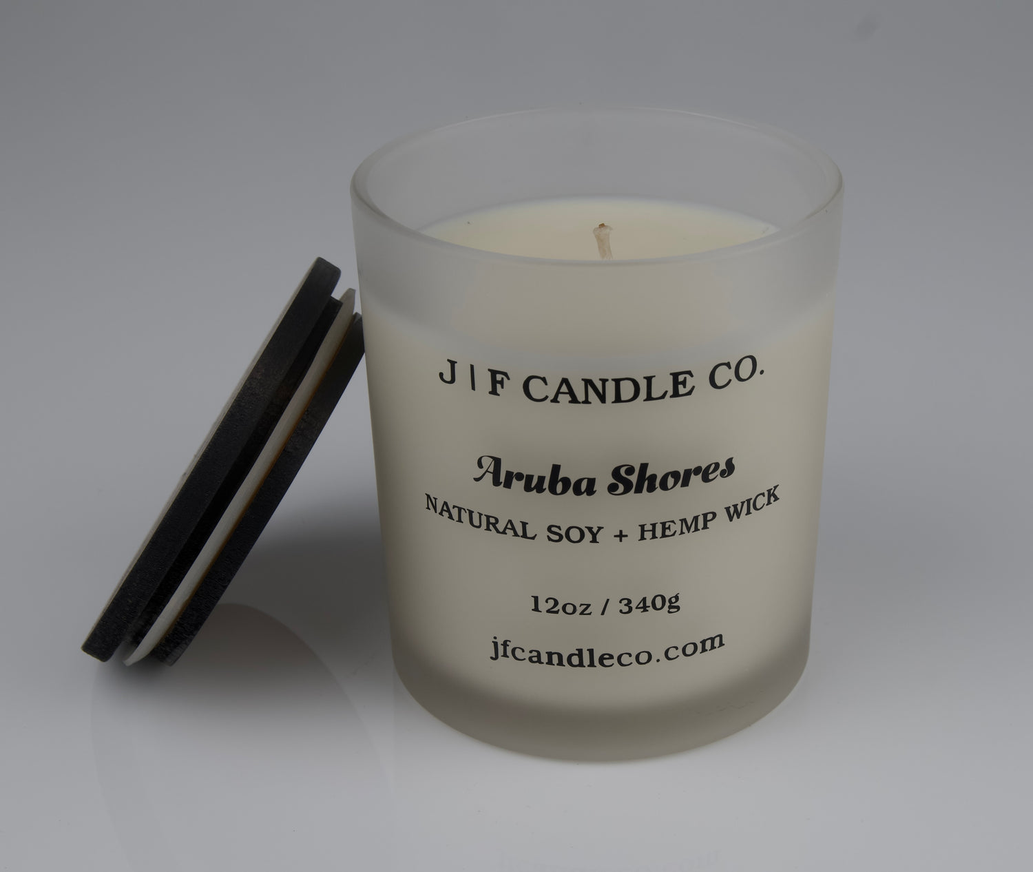 Frosted candle jar with wooden lid resting on side labeled 'J/F Candle Co. Aruba Shores'
