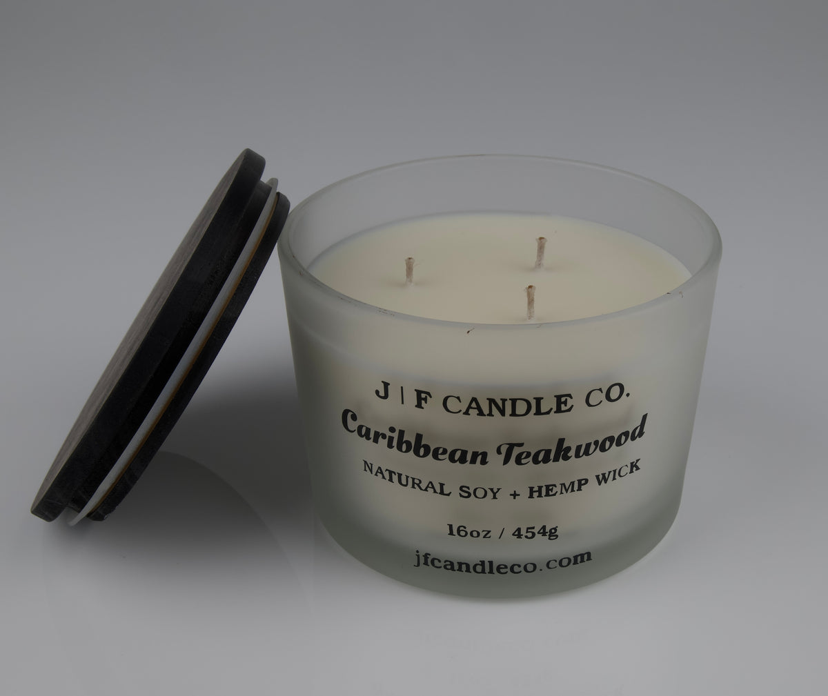 16oz Frosted glass candle with wooden lid labeled 'J|F Candle Co. Caribbean Teakwood' with lid rested on side of jar