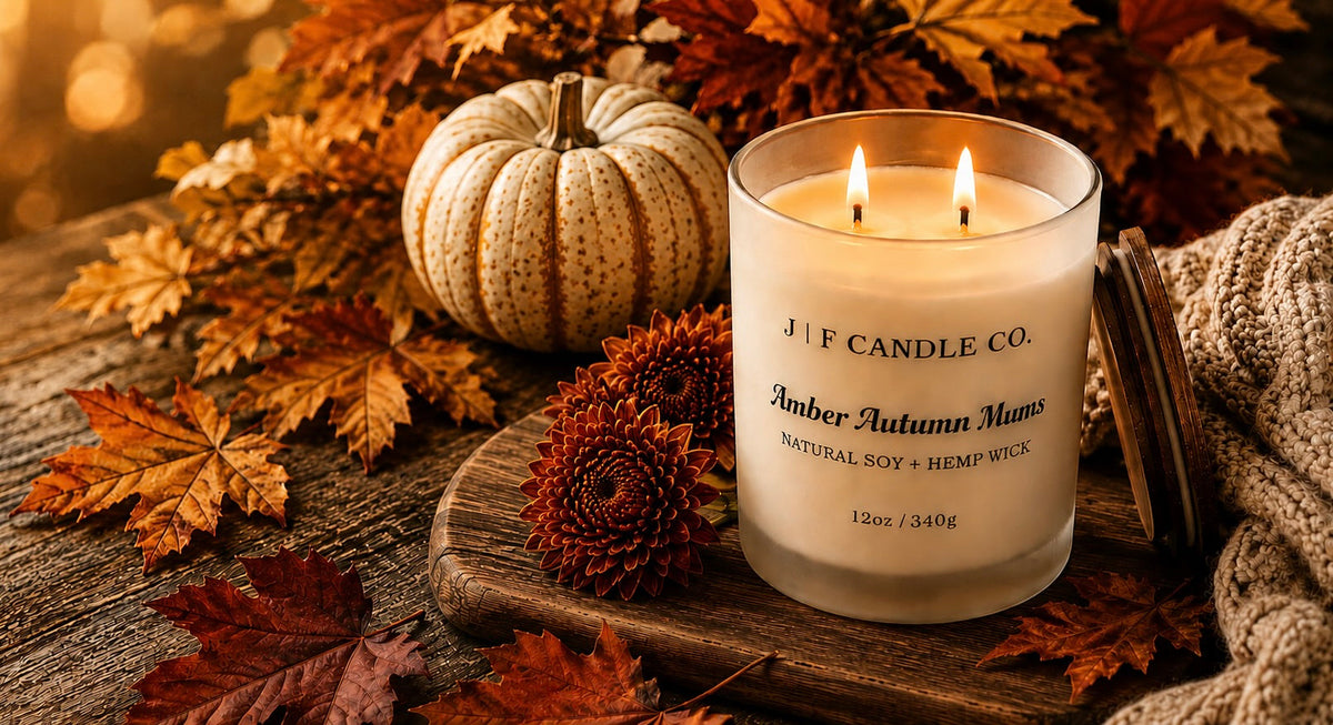 Hand-poured Amber Autumn Mums soy candle by J | F Candle Co. surrounded by autumn leaves, a white pumpkin, and fall florals on a rustic wooden table.