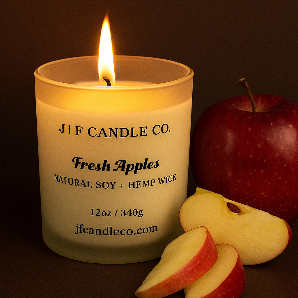A lit “Fresh Apples” candle by J | F Candle Co., crafted with natural soy and a hemp wick, glows warmly beside a whole red apple and freshly sliced apple pieces, set against a deep brown background.