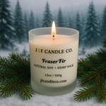 A lit “Fraser Fir” scented candle by J | F Candle Co., made with natural soy and a hemp wick, sits nestled among pine branches on a snowy forest floor. A soft, warm flame flickers against a blurred backdrop of snow-covered evergreen trees.