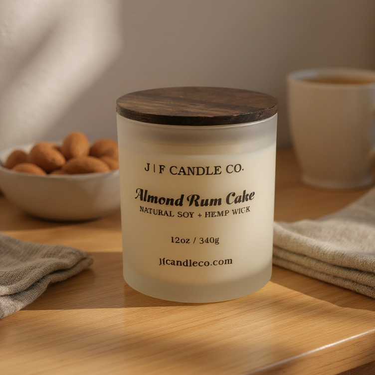 A frosted glass candle labeled “Almond Rum Cake” from J | F Candle Co. sits on a light wood surface, topped with a dark wooden lid. The candle label reads “Natural Soy + Hemp Wick – 12oz / 340g – jfcandleco.com.” In the background, there’s a bowl of almonds, a cup of coffee, and a soft beige cloth, all softly lit by natural sunlight streaming through a nearby window, creating a cozy and inviting atmosphere.