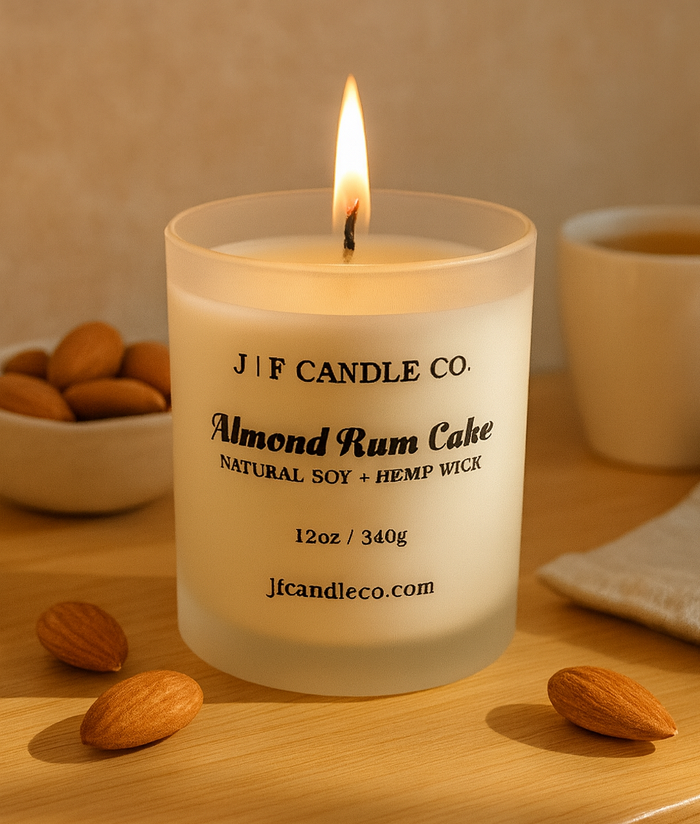 A lit almond rum cake-scented soy candle from J | F Candle Co. in a frosted glass jar sits on a light wooden surface. The candle's black text label is clearly visible, and it is surrounded by a small white bowl of almonds, a ceramic mug with a warm drink, and a folded beige cloth, all bathed in soft, natural sunlight.