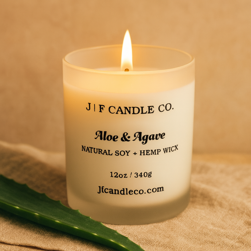 A frosted glass candle labeled 'Aloe & Agave' from J | F Candle Co. sits on a warm beige linen surface. The wick is lit with a soft, warm flame, and a green aloe leaf rests beside the candle, creating a natural, calming, spa-like atmosphere.