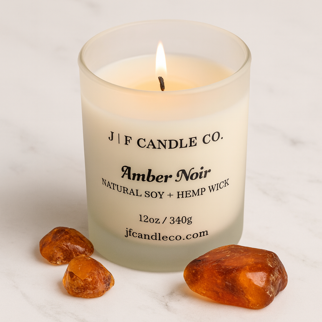 A lit Amber Noir candle from J | F Candle Co. sits on a polished white marble surface, surrounded by amber stones. The frosted glass jar displays a black serif label with the scent name and product details, while a small flame glows softly above the creamy soy wax.