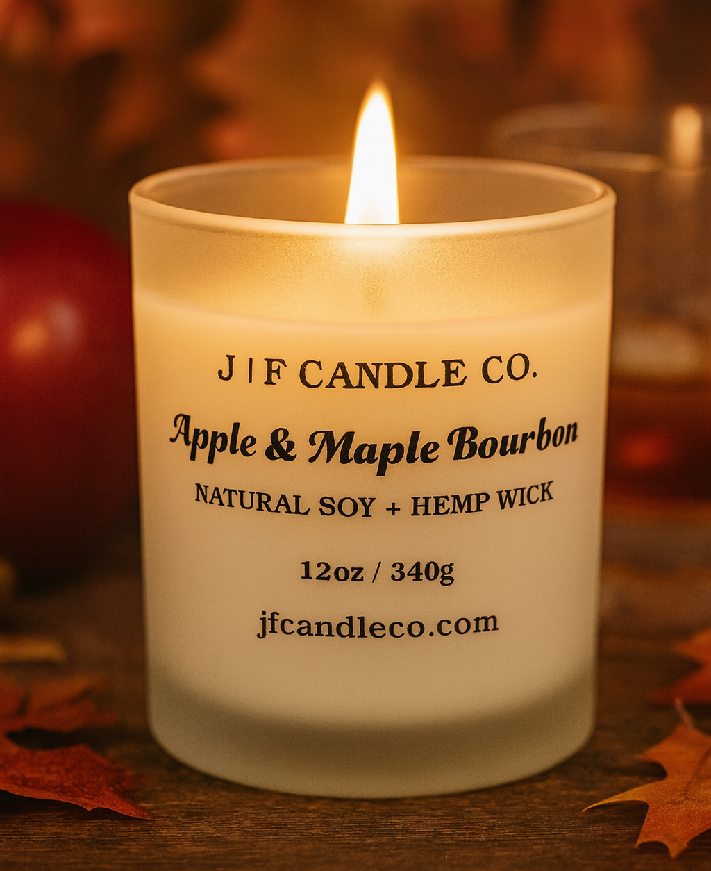 A lit Apple & Maple Bourbon soy candle by J | F Candle Co. in a frosted glass jar with the lid removed, styled in an autumn setting with apple slices and bourbon-themed props, showcasing a soft flame and original product label.