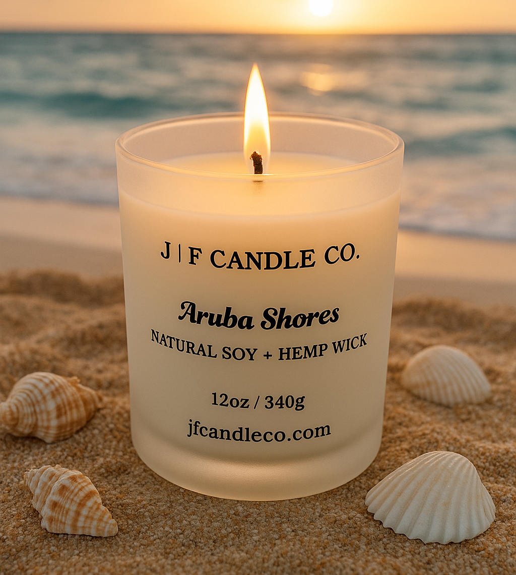 A lit "Aruba Shores" candle from J | F Candle Co. rests on a sun-kissed beach in Aruba, surrounded by seashells and coral. The frosted white glass jar glows warmly in the golden hour light, with swaying palm trees and turquoise ocean waves softly blurred in the background.
