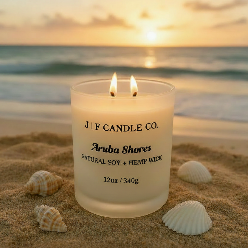 A lit "Aruba Shores" candle from J | F Candle Co. rests on a sun-kissed beach in Aruba, surrounded by seashells and coral. The frosted white glass jar glows warmly in the golden hour light, with swaying palm trees and turquoise ocean waves softly blurred in the background.