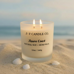 Azure Coast candle by J | F Candle Co. in a frosted glass jar with black text labeling, shown with a hemp wick. The candle is placed on light beach sand with small white seashells nearby, and a soft ocean backdrop creating a calm coastal atmosphere.