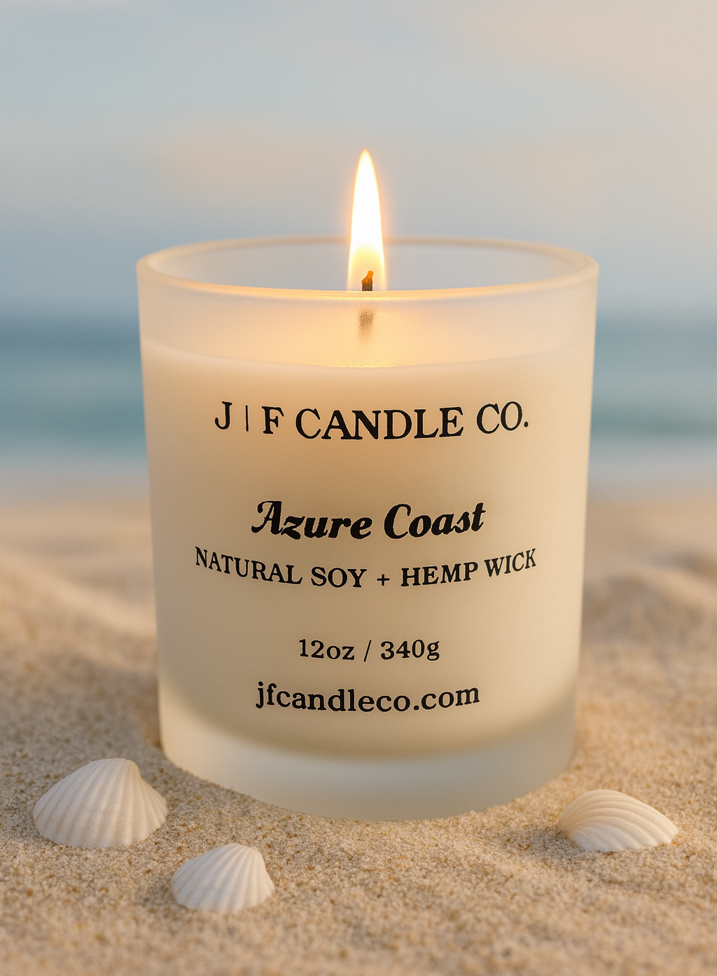 Azure Coast candle by J | F Candle Co. in a frosted glass jar with black text labeling, shown with a hemp wick. The candle is placed on light beach sand with small white seashells nearby, and a soft ocean backdrop creating a calm coastal atmosphere.