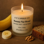 A lit "Banana Nut Bread" candle from J | F Candle Co. in a frosted glass jar sits on a wooden surface. The black label features the scent name, product size, and company website. Surrounding the candle are a ripe banana, a banana slice, walnut pieces, and a slice of banana nut bread, creating a cozy and aromatic presentation.
