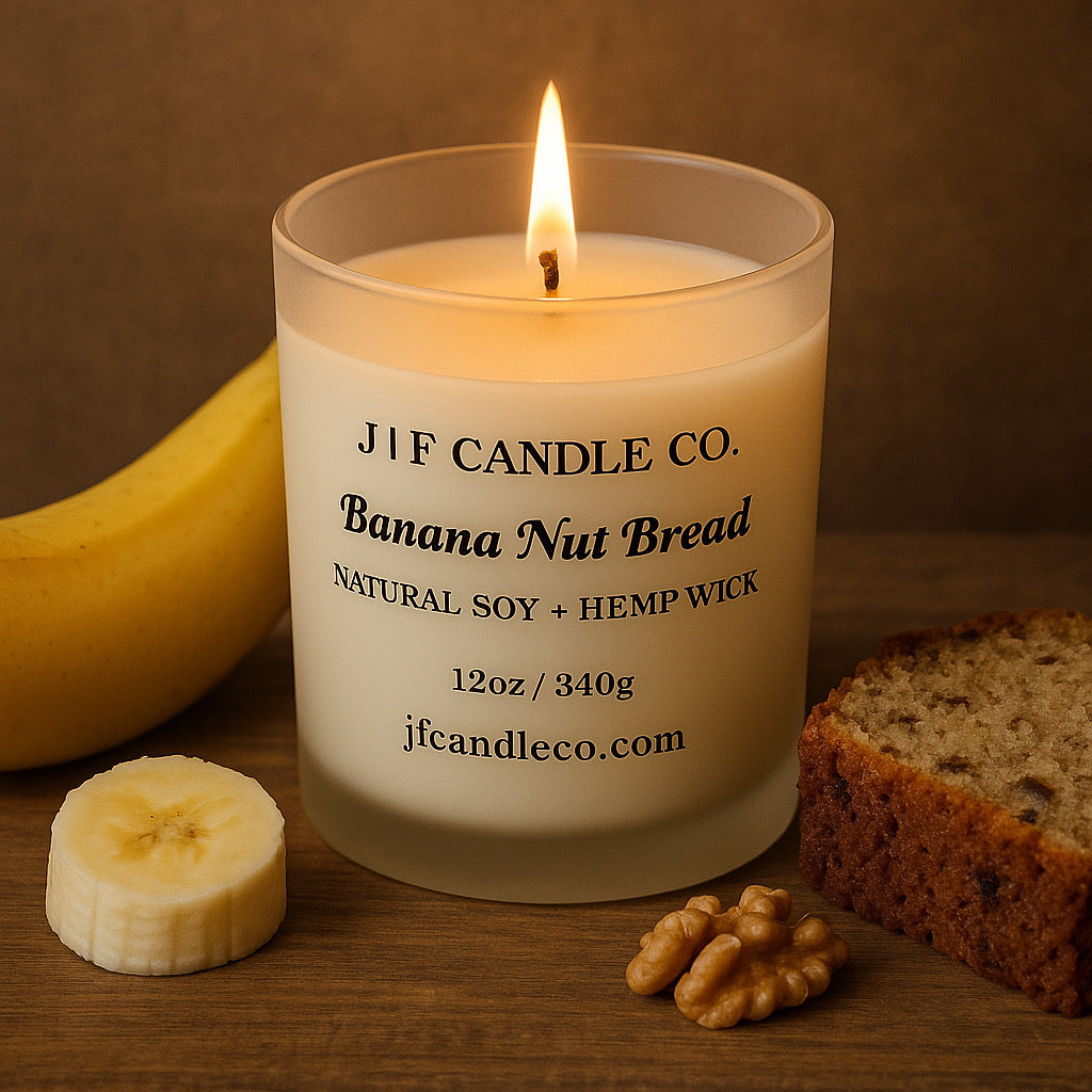 A lit "Banana Nut Bread" candle from J | F Candle Co. in a frosted glass jar sits on a wooden surface. The black label features the scent name, product size, and company website. Surrounding the candle are a ripe banana, a banana slice, walnut pieces, and a slice of banana nut bread, creating a cozy and aromatic presentation.