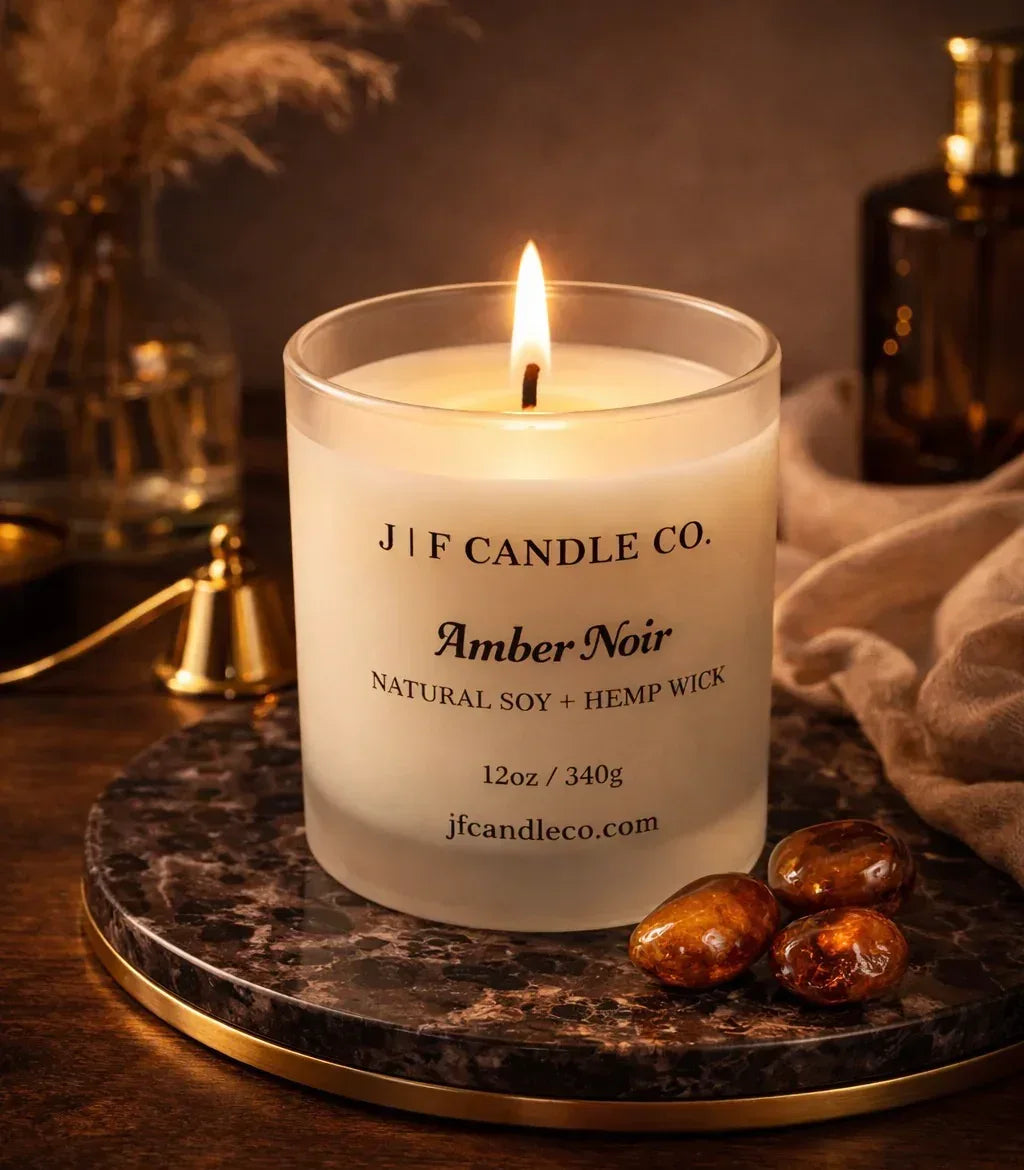 Lit Amber Noir soy candle by J | F Candle Co. in a frosted glass jar styled on marble with dark amber stones