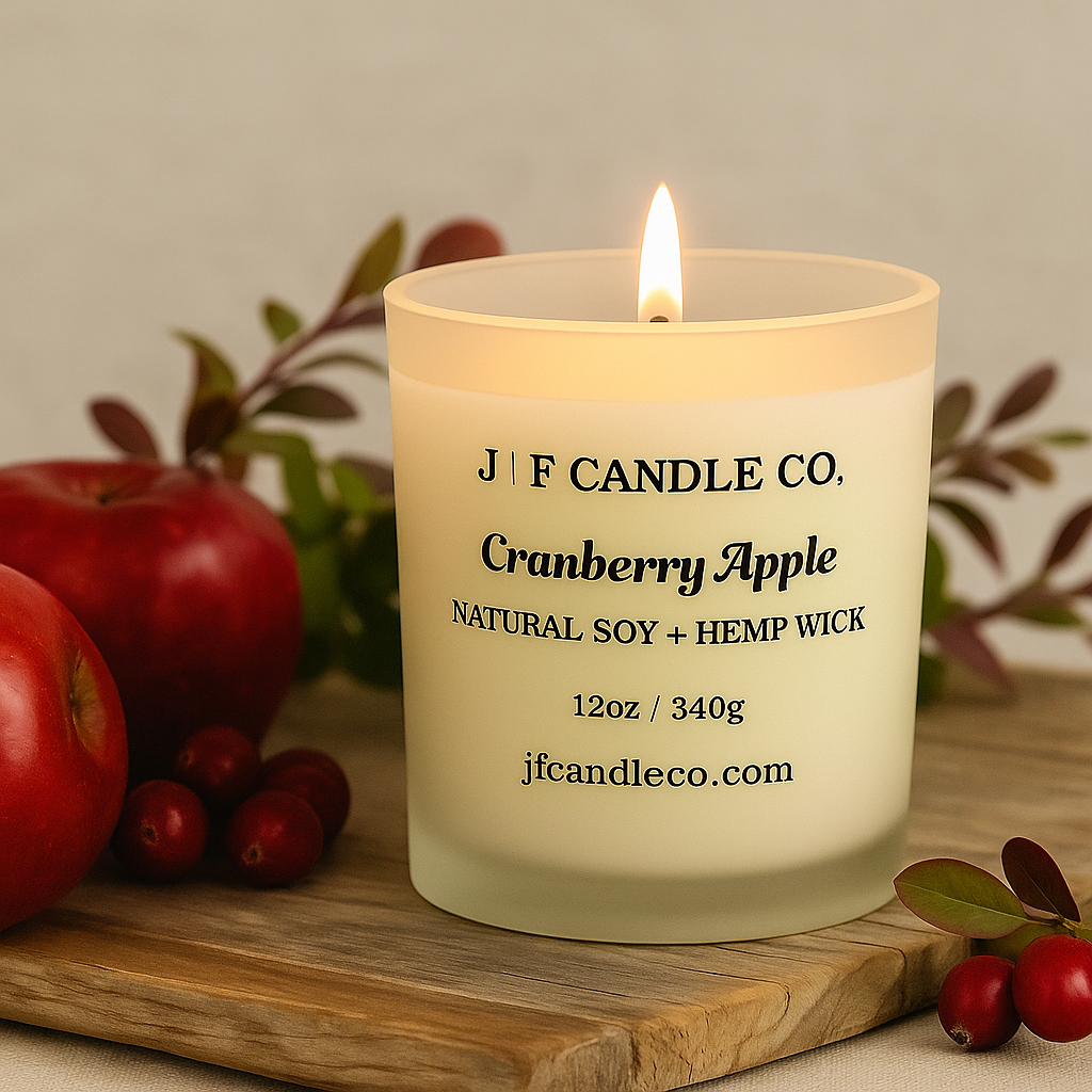 Lit Cranberry Apple soy candle by J | F Candle Co. in a frosted glass jar, styled on a rustic wood surface with fresh apples, cranberries, and green foliage — eco-luxury candle for cozy seasonal ambiance.