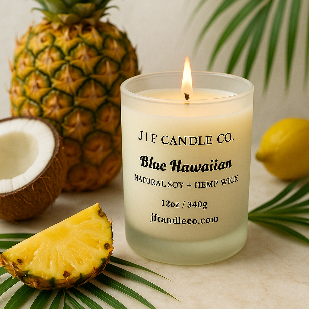 A lit Blue Hawaiian scented candle in a frosted glass jar from J | F Candle Co., styled on a light stone surface with fresh pineapple, coconut, and tropical palm leaves, in bright natural lighting.