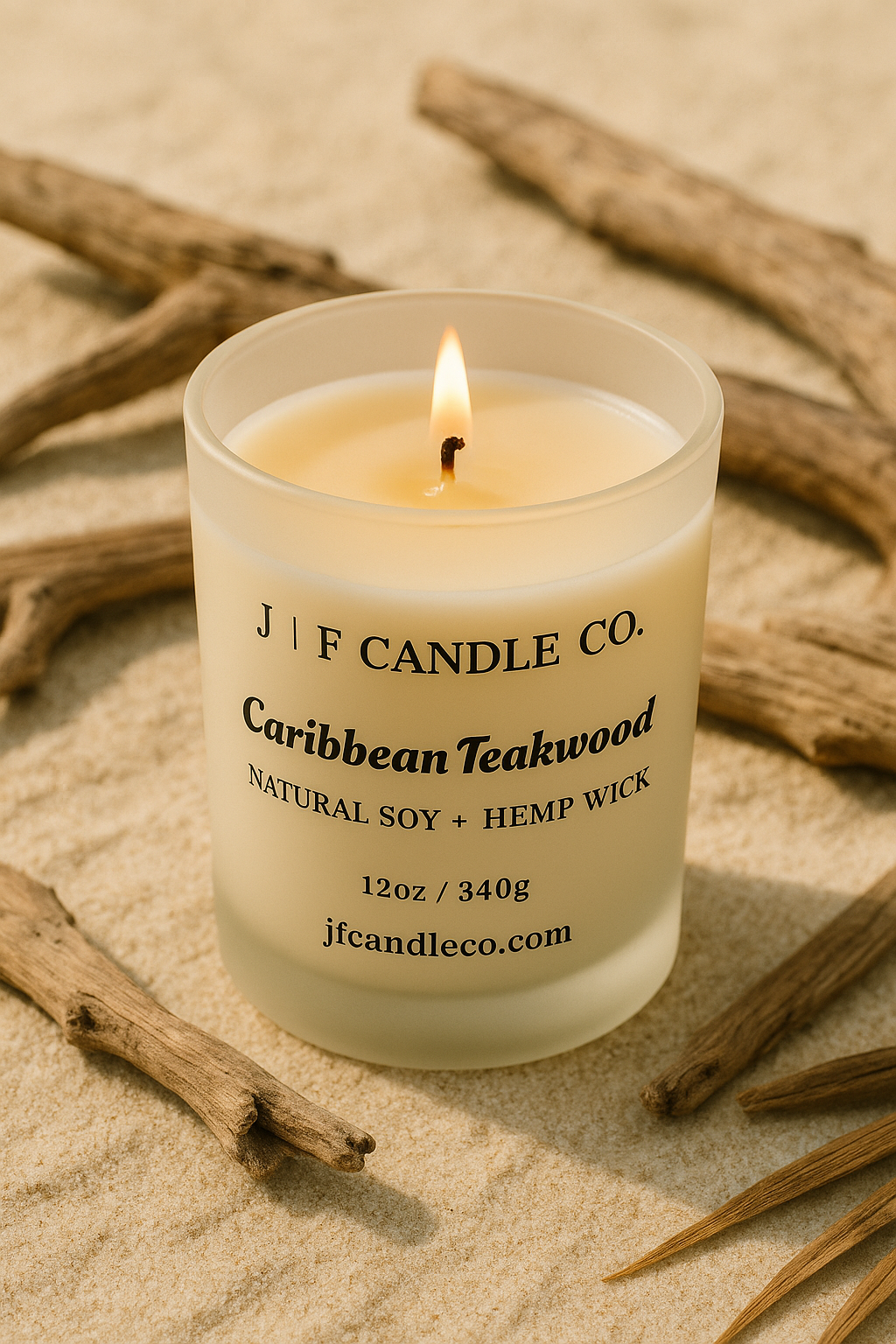 A lit Caribbean Teakwood candle in a frosted glass jar from J | F Candle Co., displayed on warm sand with pieces of natural driftwood around it, photographed in soft golden light for a coastal, earthy ambiance.