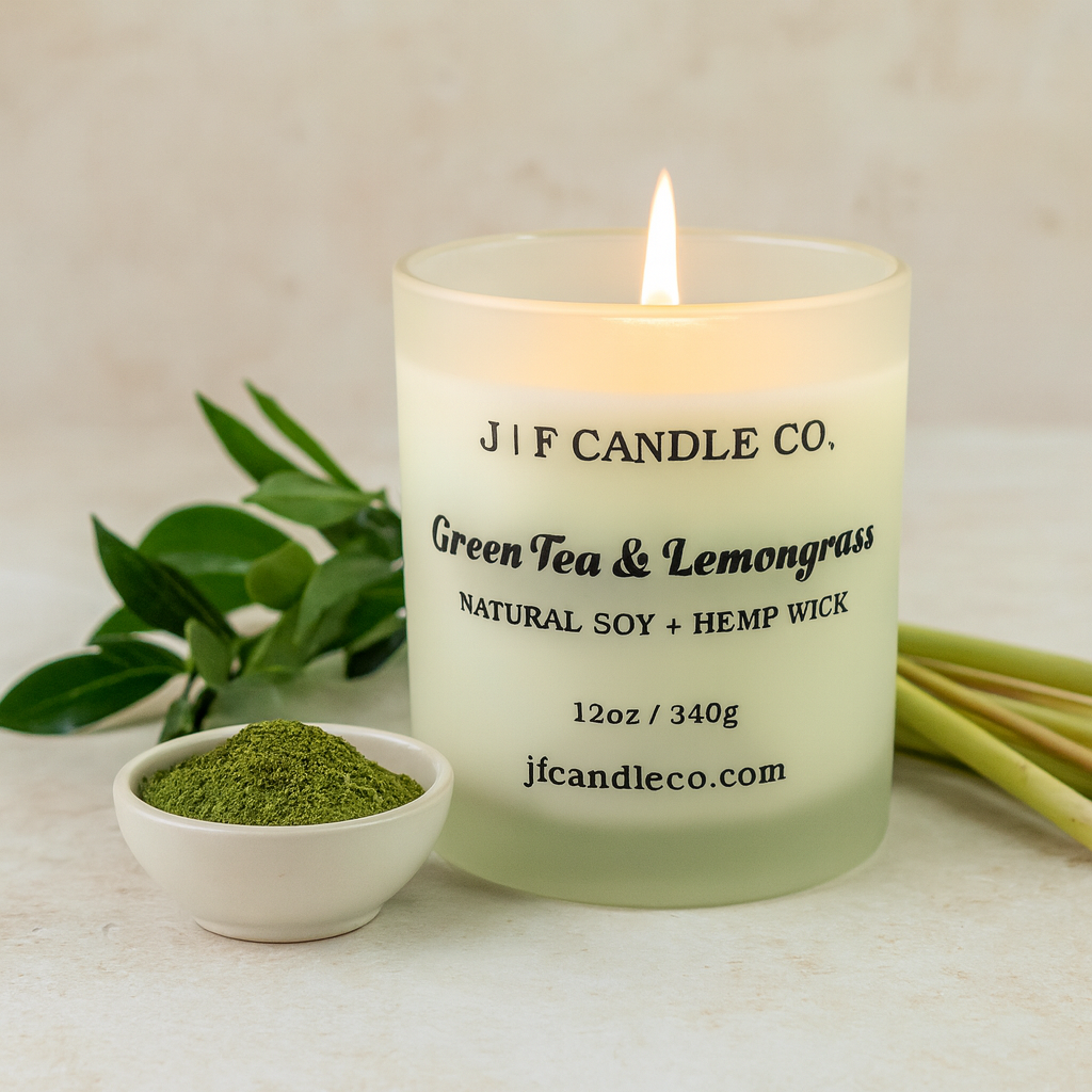 A lit J | F Candle Co. “Green Tea & Lemongrass” natural soy candle with a hemp wick glows warmly in a frosted glass jar. In front of it sits a small white bowl filled with ground green tea powder, surrounded by lemongrass stalks and fresh green leaves on a light beige stone surface under soft natural lighting.