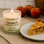A lit J | F Candle Co. “Hot Apple Pie” natural soy candle with a hemp wick glows warmly on a beige kitchen countertop beside a white plate holding a slice of apple pie. Several fresh red apples are softly blurred in the background, creating a cozy, home-baked autumn atmosphere.