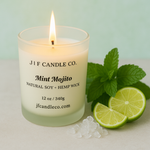 A lit Mint Mojito candle from J | F Candle Co. in a frosted glass jar sits on a light surface surrounded by fresh mint leaves, lime slices, and crushed ice, creating a bright and refreshing tropical aesthetic.