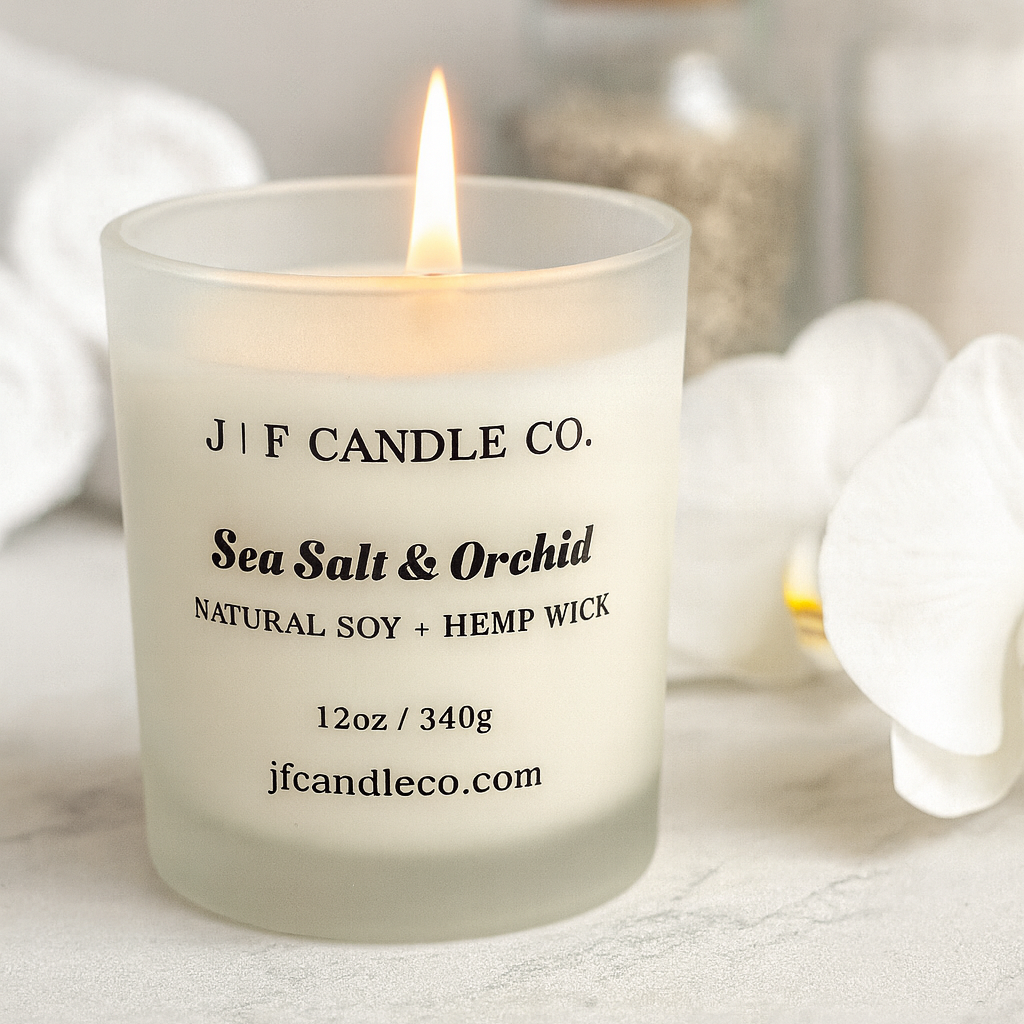 Lit J | F Candle Co. Sea Salt & Orchid candle in a frosted glass jar, styled with white spa towels, smooth stones, and green leaves on a soft neutral background — clean, calming luxury soy candle scene.
