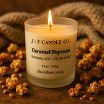 A frosted glass jar candle labeled “Caramel Popcorn” from J | F Candle Co. sits on a wooden surface. The wick is lit, casting a warm golden light. Caramel-coated popcorn pieces are scattered around the candle, with a textured beige blanket softly blurred in the background.