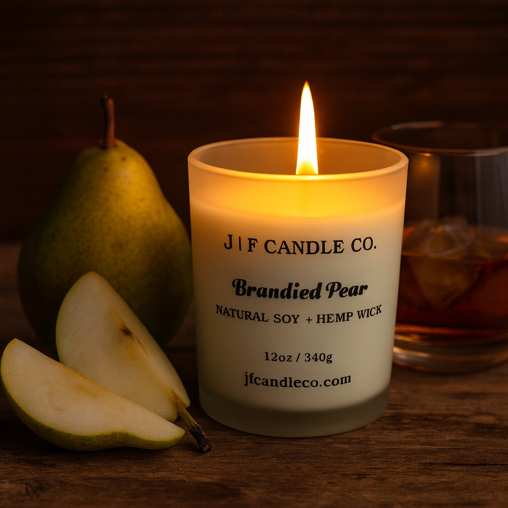 A lit Brandied Pear candle by J | F Candle Co. in a frosted glass jar on a wooden surface, surrounded by fresh pear slices and a glass of brandy in the background, creating a warm, cozy atmosphere.