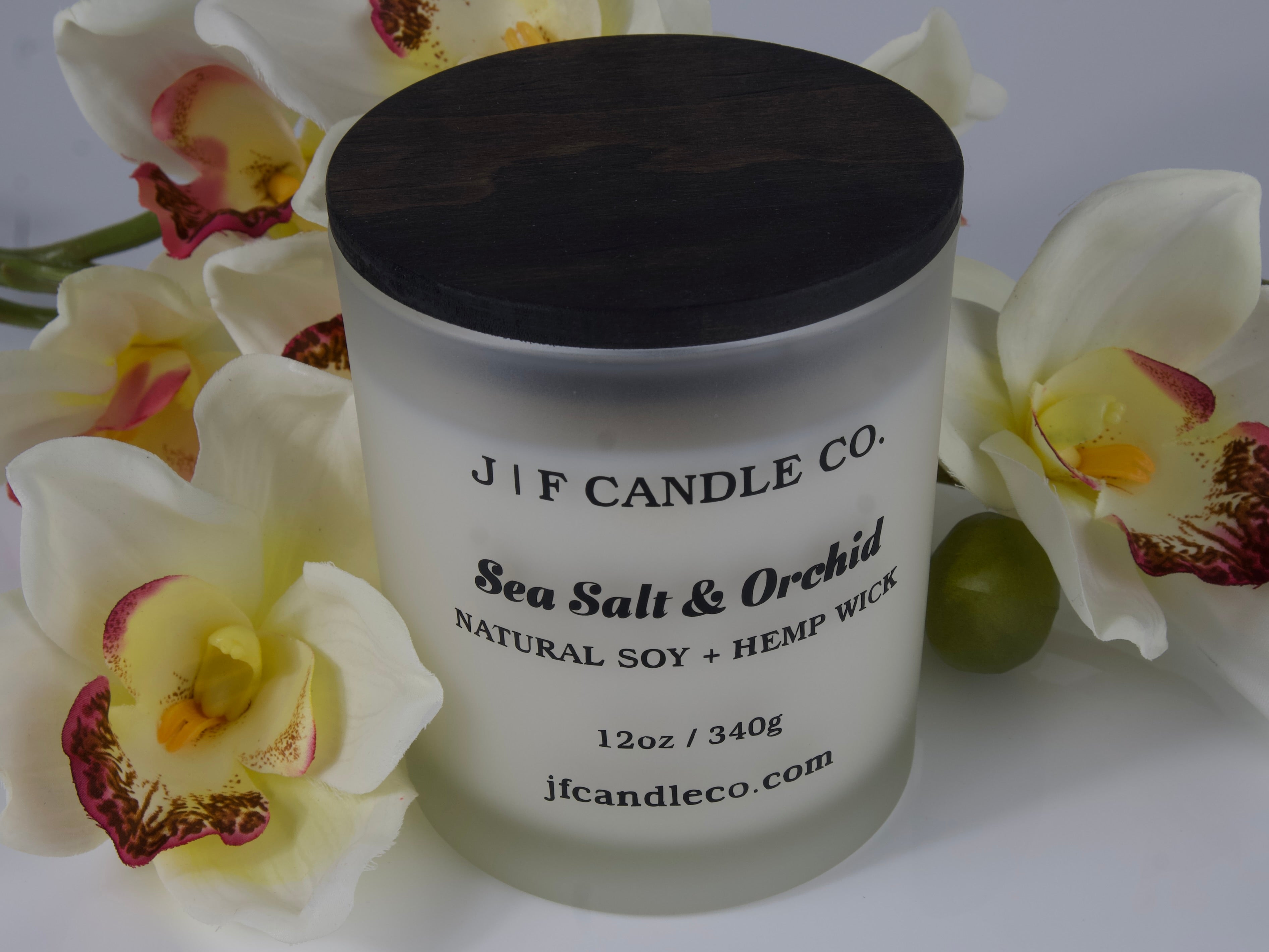ChatGPT said:
A frosted glass candle with a dark wooden lid labeled "J | F Candle Co. Sea Salt & Orchid, Natural Soy + Hemp Wick, 12oz / 340g, jfcandleco.com" is surrounded by white orchids with pink and yellow centers. The elegant floral arrangement complements the candle's clean, sophisticated design.