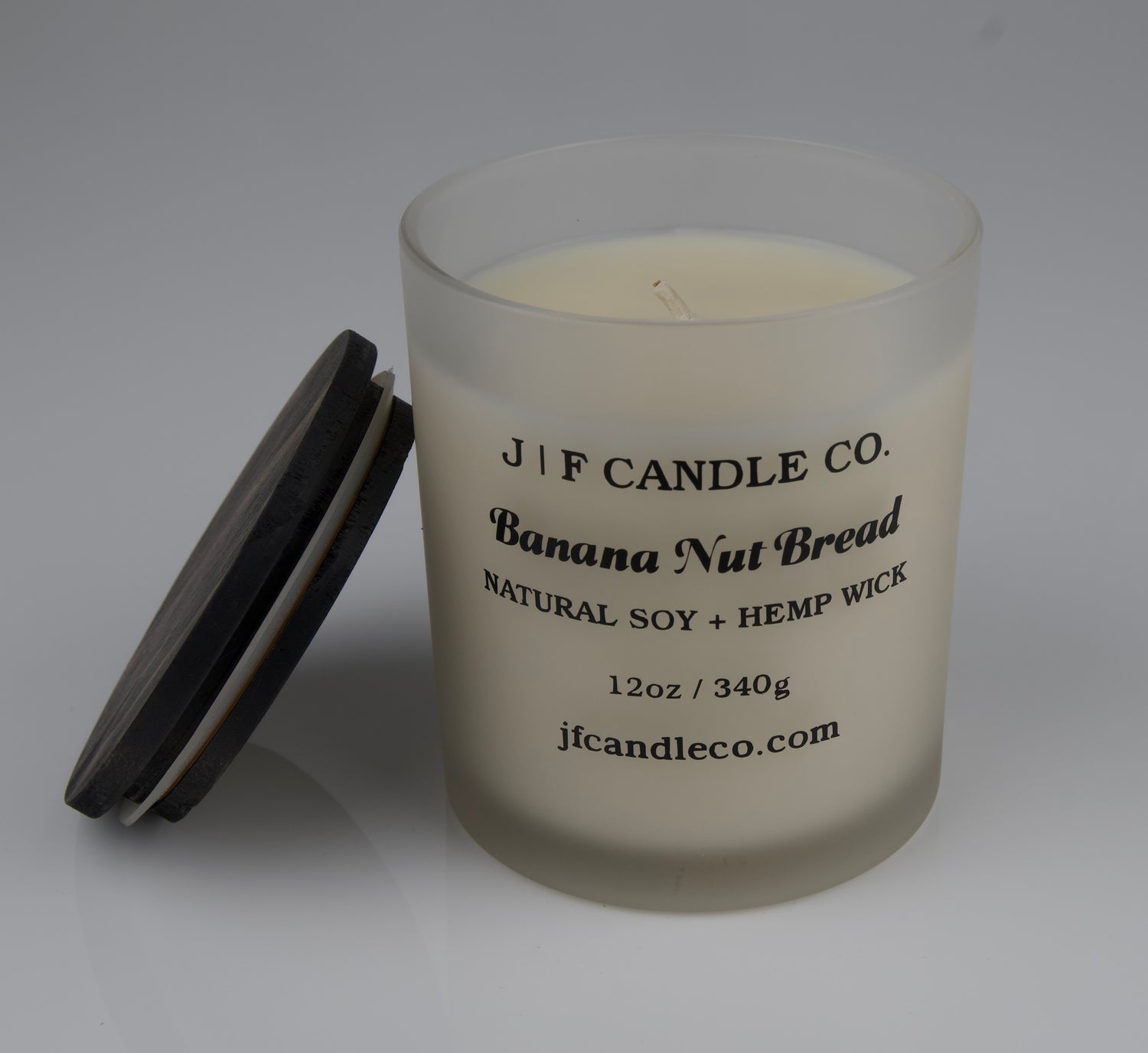 JF Candle Co. 12oz Banana Nut Bread soy candle with single hemp wick in frosted glass jar, dark wooden lid resting beside it.