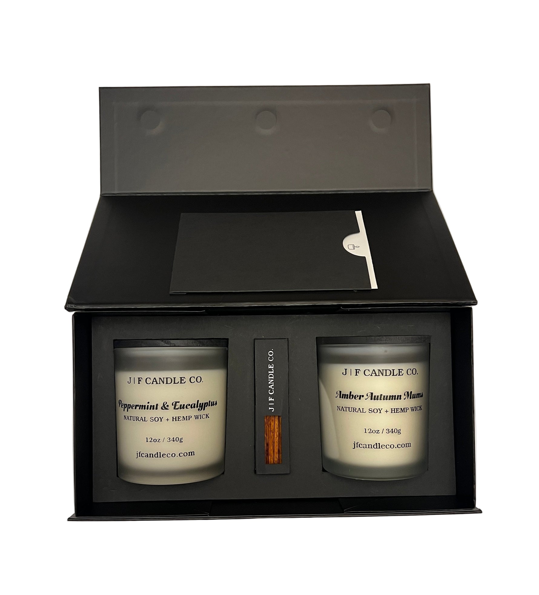 An open black gift box from J | F Candle Co. contains two frosted glass soy candles labeled “Peppermint & Eucalyptus” and “Amber Autumn Mums,” with a box of matches placed neatly in the center. The box has a clean matte finish and a minimalist insert card, photographed against a plain white background.