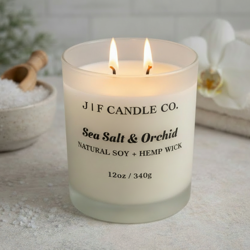 A lit Sea Salt & Orchid candle from J | F Candle Co. sits on a light stone surface, surrounded by a ceramic bowl of coarse sea salt with a wooden scoop, a white orchid flower, and soft background greenery. The frosted glass jar glows warmly, highlighting the black label text and creating a calm, spa-like ambiance.
