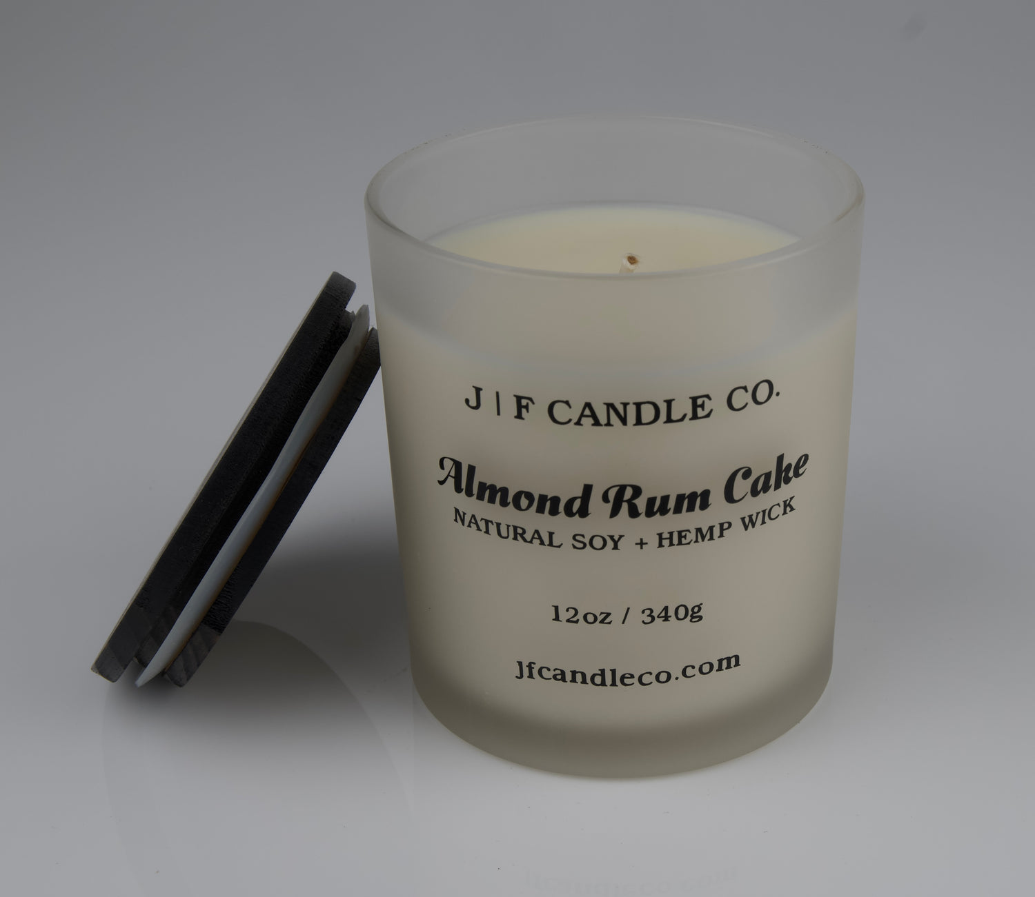JF Candle Co. 12oz Almond Rum Cake soy candle with single hemp wick in frosted glass jar, dark wooden lid resting beside it.