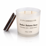 J | F Candle Co. Amber Autumn Mums luxury soy candle in a frosted glass 12 oz jar with double unlit hemp wicks, black label, wooden lid beside jar, and soft reflection on a clean white background.