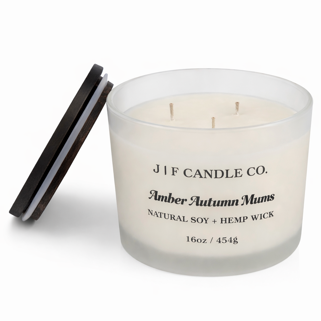 J | F Candle Co. Amber Autumn Mums 16 oz luxury soy candle in a frosted glass jar with three unlit hemp wicks, black label, dark wooden lid resting beside the jar, and clean white background.