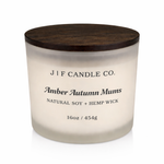 Amber Autumn Mums luxury soy candle in a frosted glass jar with wooden lid, softly lit on a clean white background with a subtle reflection beneath the base.