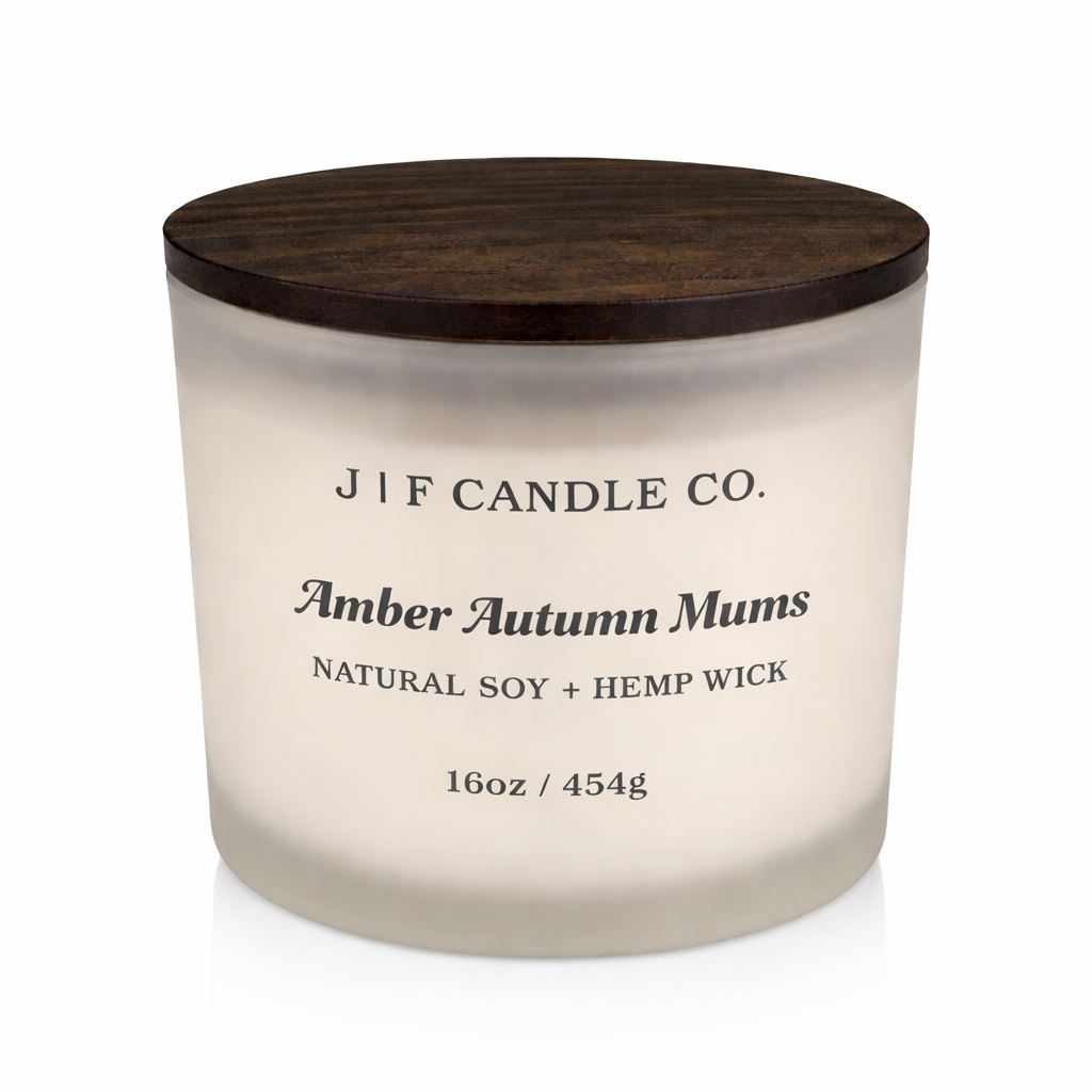 Amber Autumn Mums luxury soy candle in a frosted glass jar with wooden lid, softly lit on a clean white background with a subtle reflection beneath the base.