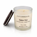 Amber Noir soy candle by J | F Candle Co. in a frosted glass jar with wooden lid resting beside it, featuring a clean white background and a soft reflective surface for a refined, luxury product presentation.