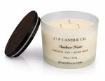 Amber Noir luxury soy candle in a frosted glass jar with wooden lid beside it, featuring three wicks and a soft reflective surface on a clean white background.