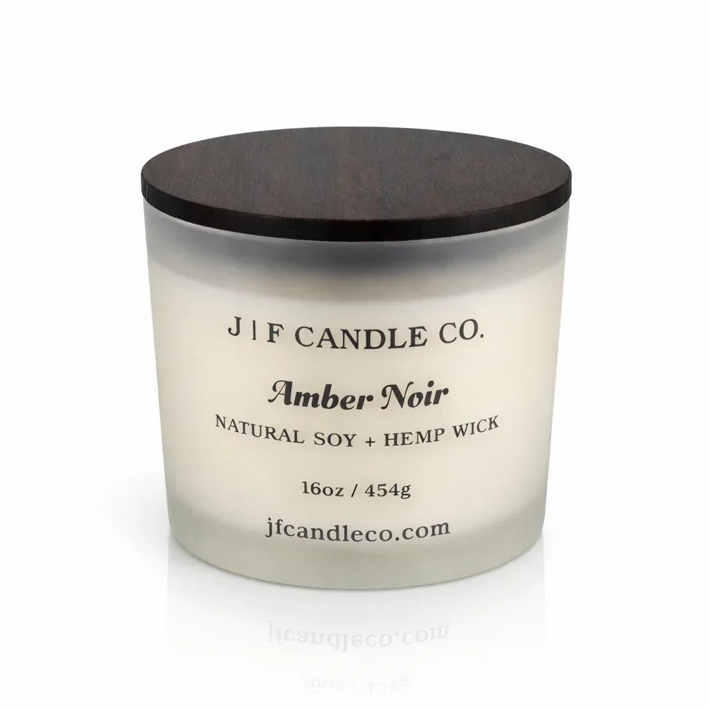 Luxury frosted glass soy candle with dark wooden lid labeled “Amber Noir,” softly illuminated on a clean white background with a subtle reflection beneath the jar.