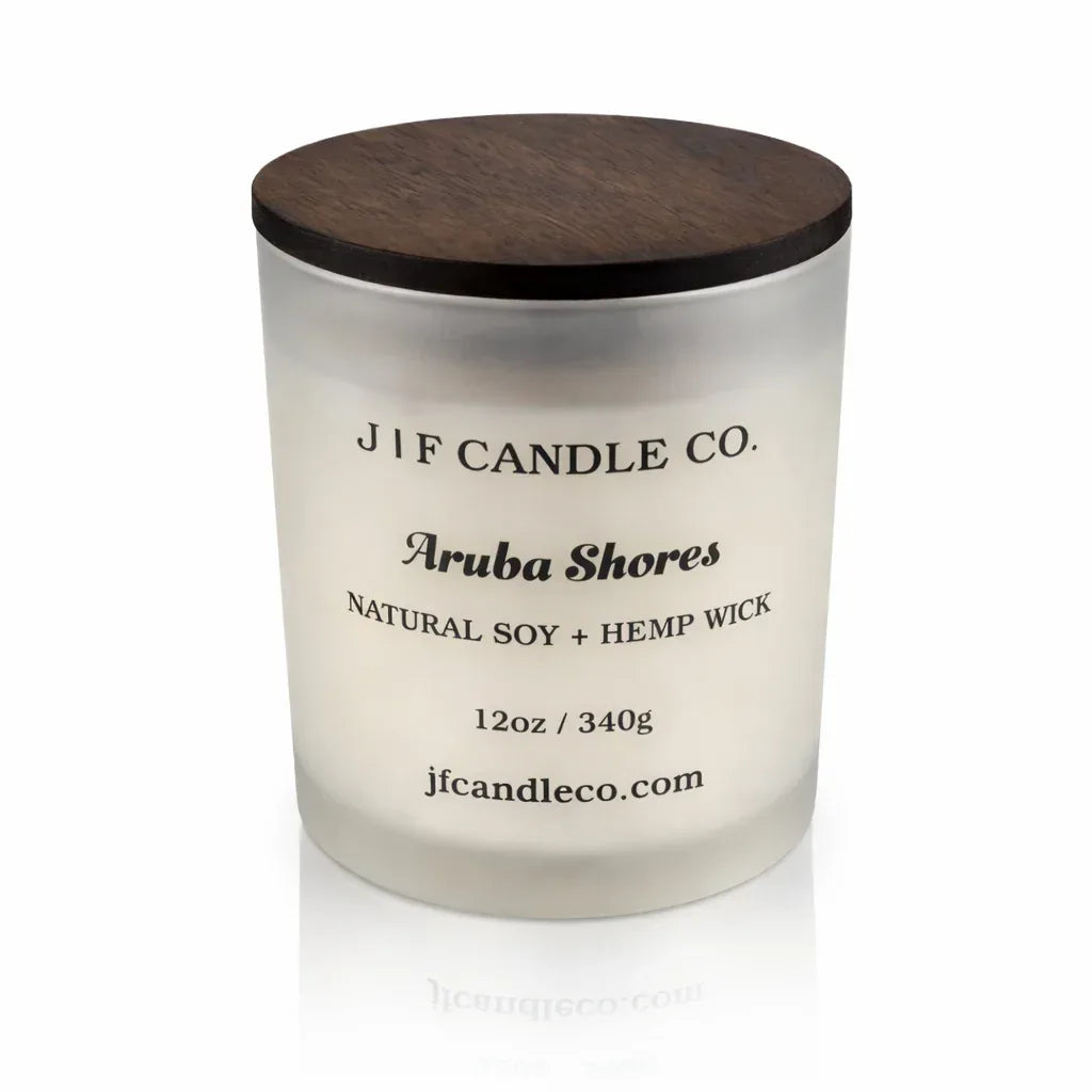 Frosted glass J | F Candle Co. Aruba Shores soy candle with dark wooden lid, softly reflected on a clean white surface for a minimalist luxury product presentation.
