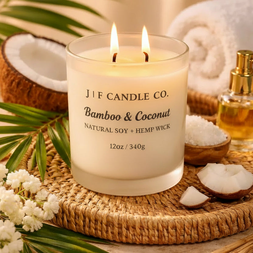 Luxury J | F Candle Co. Bamboo & Coconut soy candle with a lit wick in a frosted glass jar, styled on a woven tray with coconut pieces, tropical palm leaves, spa towel, and warm ambient lighting.