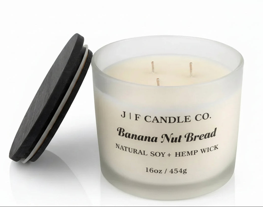 J F Candle Co Banana Nut Bread 16 oz soy candle with three wicks in a frosted glass jar with wooden lid beside it on a clean white background with soft reflection