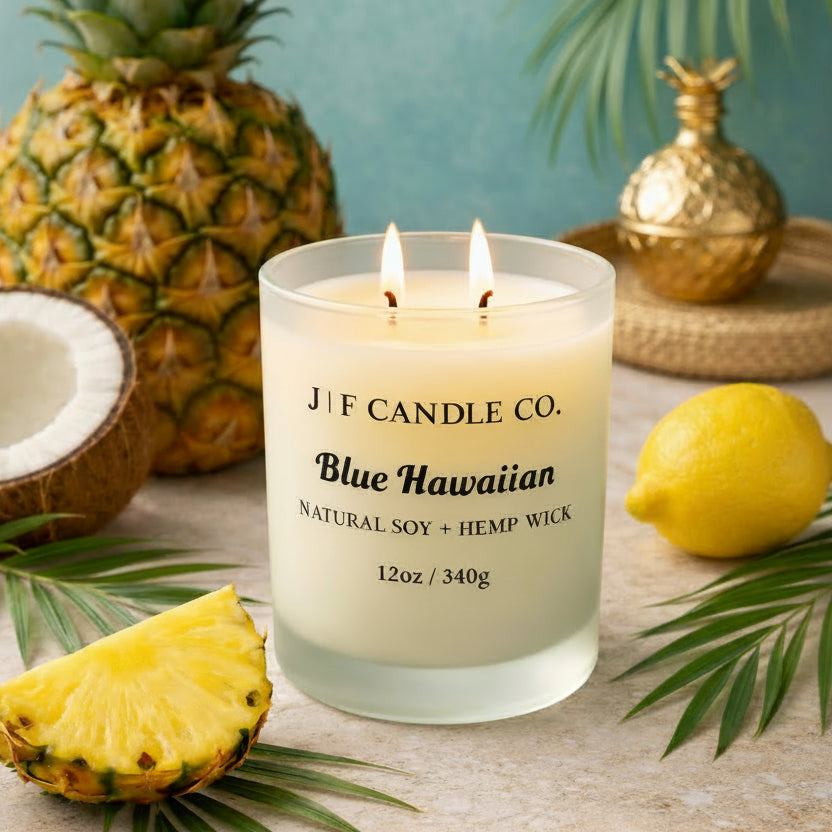 Blue Hawaiian luxury soy candle by J | F Candle Co. in a frosted glass jar surrounded by pineapple, coconut, and tropical leaves.