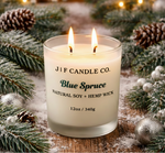 J | F Candle Co. Blue Spruce soy candle with a lit wick in a frosted glass jar, styled on a wooden surface with blue spruce branches, pinecones, and soft snow accents in a warm holiday setting
