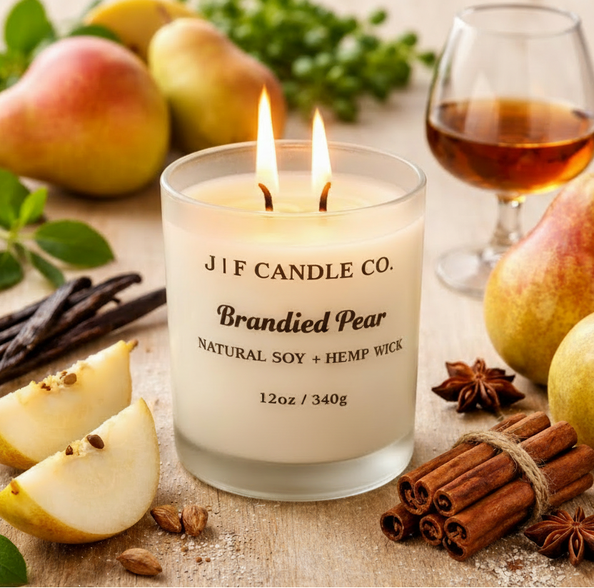 J | F Candle Co. Brandied Pear soy candle with a lit wick in a frosted glass jar, styled on a light wood surface with fresh pears, vanilla beans, cinnamon sticks, and a glass of brandy in a warm, upscale kitchen setting