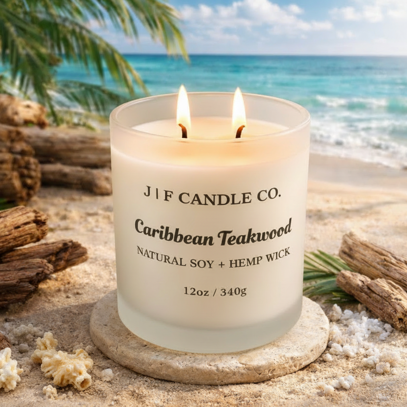 J | F Candle Co. Caribbean Teakwood 12 oz natural soy candle with lit wick in frosted glass jar styled on a beach with driftwood, sand, and ocean background
